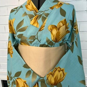 May include: A light blue kimono featuring a repeating pattern of large golden-yellow tulip-like flowers and dark olive green leaves. The garment is styled with a wide, solid beige obi sash wrapped around the midsection, displayed on a mannequin.