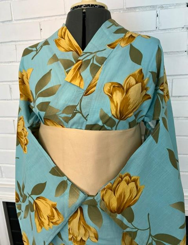 May include: A light blue kimono featuring a repeating pattern of large golden-yellow tulip-like flowers and dark olive green leaves. The garment is styled with a wide, solid beige obi sash wrapped around the midsection, displayed on a mannequin.