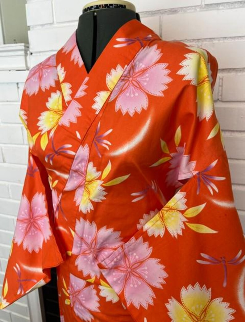 Może przedstawiać: Jaskrawopomarańczowe kimono z powtarzającym się wzorem dużych r&oacute;żowych i ż&oacute;łtych kwiat&oacute;w wiśni przeplatanych małymi fioletowymi ważkami. Odzież ma tradycyjny kr&oacute;j kopertowy z szerokimi rękawami i usztywnianym kołnierzem.