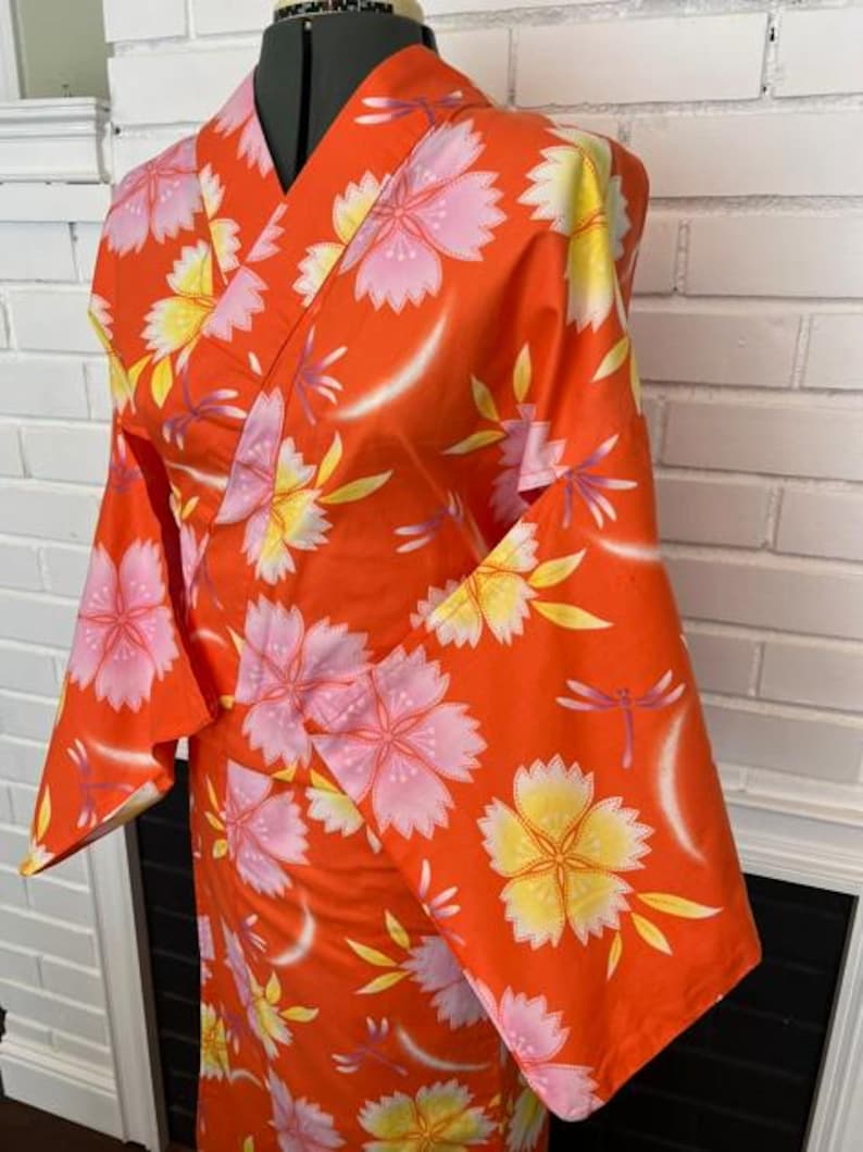 Może przedstawiać: Jaskrawopomarańczowe kimono z powtarzającym się wzorem dużych r&oacute;żowych i ż&oacute;łtych kwiat&oacute;w wiśni, przeplatanych małymi fioletowymi ważkami. Ubi&oacute;r posiada szerokie rękawy, dekolt w serek oraz kopertowy kr&oacute;j zaprezentowany na manekinie.