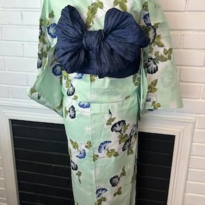 Op de afbeelding: Een licht muntgroene kimono met een herhalend patroon van blauwe en witte ochtendgloriebloemen met groene bladeren. Het kledingstuk is in de taille getailleerd met een grote, donker marineblauwe, gestructureerde obi-sjerp in een strikvorm.