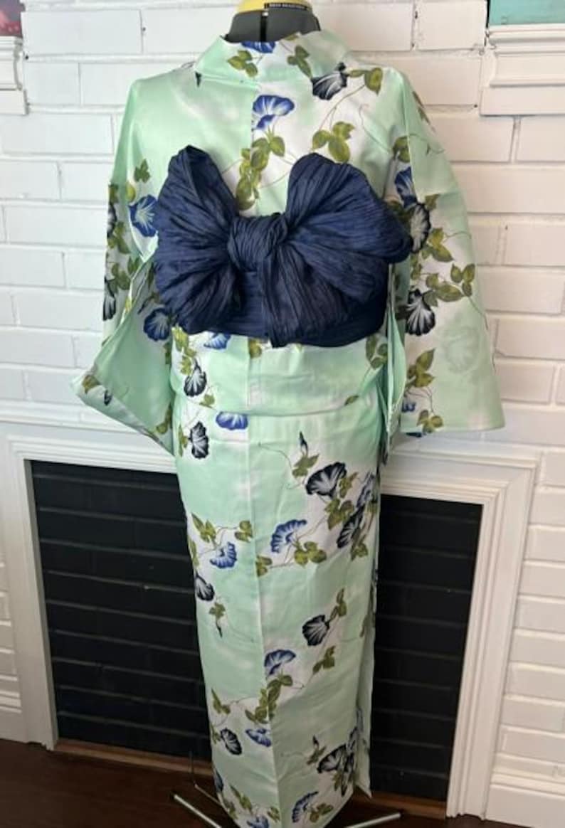 Op de afbeelding: Een licht muntgroene kimono met een herhalend patroon van blauwe en witte ochtendgloriebloemen met groene bladeren. Het kledingstuk is in de taille getailleerd met een grote, donker marineblauwe, gestructureerde obi-sjerp in een strikvorm.