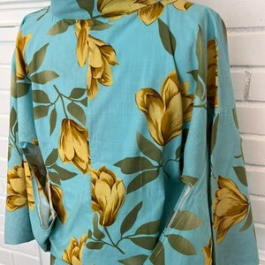 May include: A light blue kimono-style top featuring a repeating pattern of large golden-yellow flowers and dark olive green leaves. The garment has a high collar, wide sleeves, and a structured fit, displayed on a mannequin with a visible neck adjustment dial.