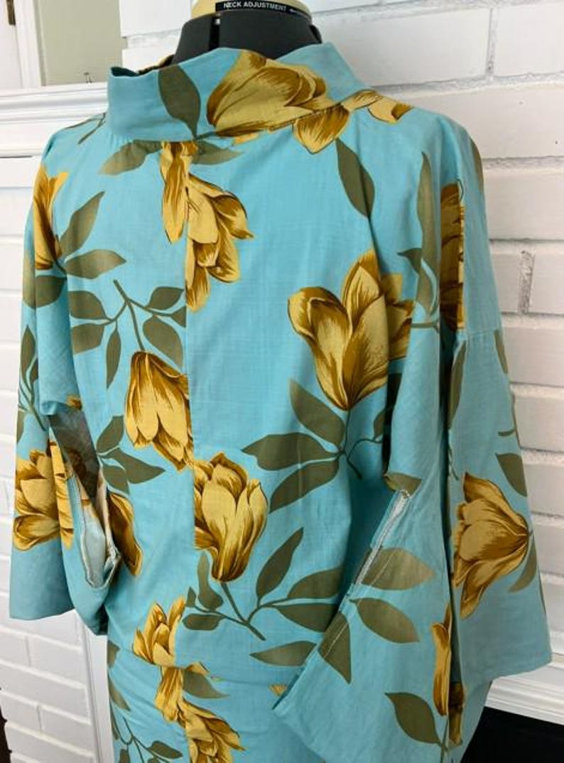 May include: A light blue kimono-style top featuring a repeating pattern of large golden-yellow flowers and dark olive green leaves. The garment has a high collar, wide sleeves, and a structured fit, displayed on a mannequin with a visible neck adjustment dial.