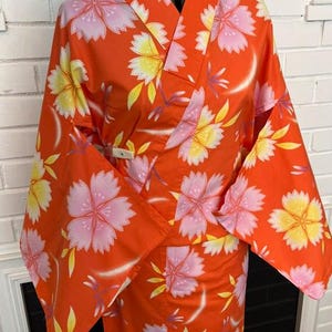 Może przedstawiać: Jaskrawopomarańczowe kimono z powtarzającym się wzorem dużych, stylizowanych kwiat&oacute;w wiśni w odcieniach jasnego r&oacute;żu i ż&oacute;łci. Ubi&oacute;r ma szerokie, luźne rękawy, tradycyjny kr&oacute;j kopertowy oraz małe, białe, prostokątne zapięcie.