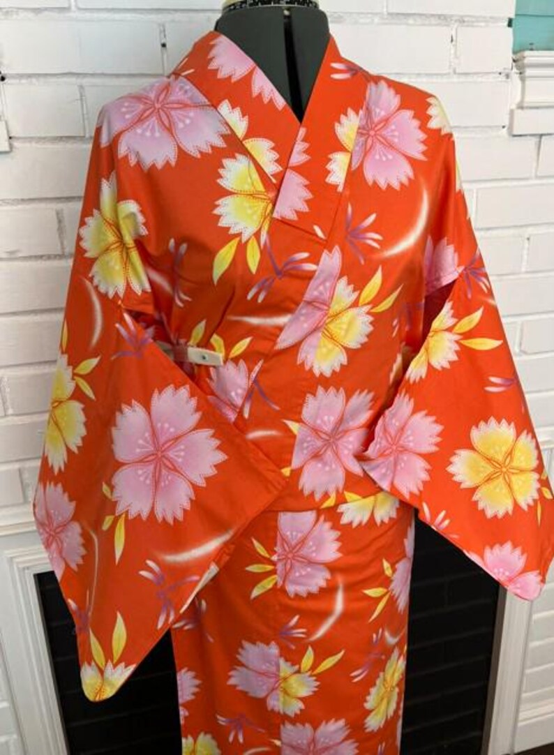 Może przedstawiać: Jaskrawopomarańczowe kimono z powtarzającym się wzorem dużych, stylizowanych kwiat&oacute;w wiśni w odcieniach jasnego r&oacute;żu i ż&oacute;łci. Ubi&oacute;r ma szerokie, luźne rękawy, tradycyjny kr&oacute;j kopertowy oraz małe, białe, prostokątne zapięcie.