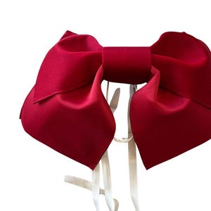 May include: A large, deep red bow with a smooth, satin-like finish. The bow features two wide, structured loops and two long, thin white ribbons hanging down from the center knot, providing a clear contrast against the vibrant red fabric.