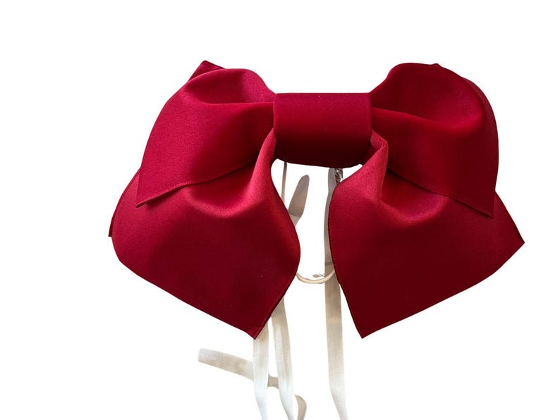 May include: A large, deep red bow with a smooth, satin-like finish. The bow features two wide, structured loops and two long, thin white ribbons hanging down from the center knot, providing a clear contrast against the vibrant red fabric.