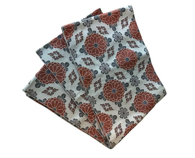 May include: A folded textile featuring a repeating pattern of rust-coloured chrysanthemum flowers, small grey floral accents, and geometric diamond shapes on a light blue background with a fine, vertical ribbed texture throughout the fabric surface.