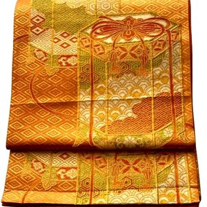 Op de afbeelding: Gevouwen oranje en gouden stof met ingewikkelde patronen. Het ontwerp bevat geometrische vormen, bloemmotieven en een centraal decoratief element met een strik en kwasten. De stof heeft een getextureerde uitstraling en een rijk kleurenpalet.