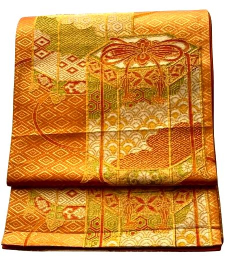 Op de afbeelding: Gevouwen oranje en gouden stof met ingewikkelde patronen. Het ontwerp bevat geometrische vormen, bloemmotieven en een centraal decoratief element met een strik en kwasten. De stof heeft een getextureerde uitstraling en een rijk kleurenpalet.