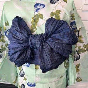 Op de afbeelding: Een licht muntgroene yukata met een bloemenpatroon van blauwe ochtendgloriebloesems en groene bladeren, in de taille gestrikt met een grote, donker marineblauwe obi-sjerp die is gevormd tot een volumineuze, gestructureerde strik op de rug.