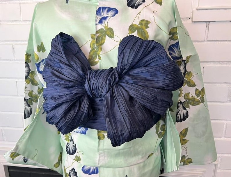 Op de afbeelding: Een licht muntgroene yukata met een bloemenpatroon van blauwe ochtendgloriebloesems en groene bladeren, in de taille gestrikt met een grote, donker marineblauwe obi-sjerp die is gevormd tot een volumineuze, gestructureerde strik op de rug.