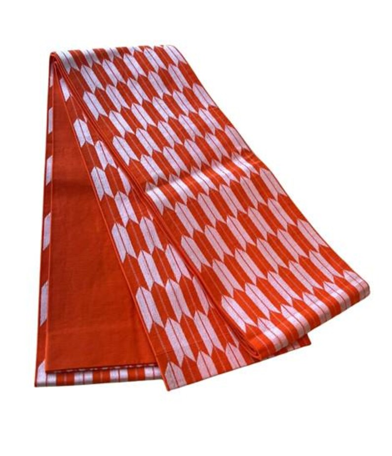 May include: A folded textile featuring a repeating pattern of white arrow-like shapes on a vibrant orange background. The fabric displays a consistent geometric design across its surface, with one section showing a solid orange color and another section displaying the arrow motif.