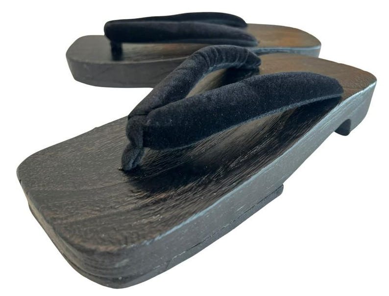 May include: A pair of traditional Japanese geta sandals featuring dark brown wooden platforms with a rectangular shape and black velvet thong straps. The wooden base shows a visible grain texture, and the elevated design includes a sturdy heel support.