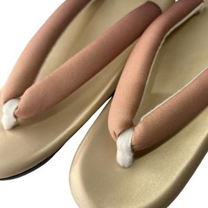 May include: A pair of traditional Japanese geta sandals. The sandals have a light gold base with a slight sheen, and the straps are a gradient of pink and brown. The thong portion is white.