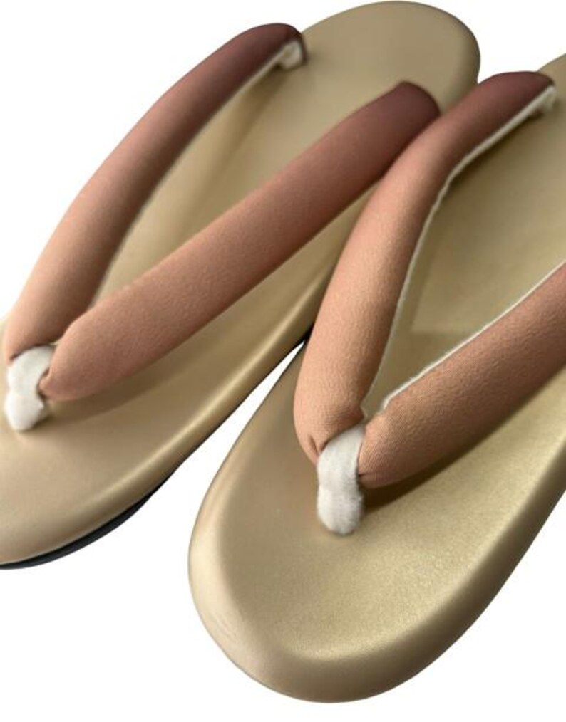 May include: A pair of traditional Japanese geta sandals. The sandals have a light gold base with a slight sheen, and the straps are a gradient of pink and brown. The thong portion is white.
