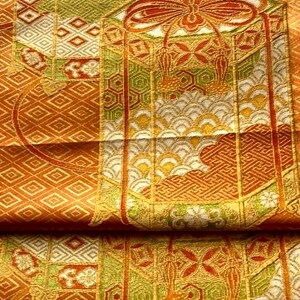 Op de afbeelding: Detail van een gevouwen textiel met een ingewikkeld patroon. Het ontwerp bevat geometrische vormen, bloemmotieven en een grote strik in oranje, goud, groen en wit. De stof lijkt zijde of een vergelijkbaar luxueus materiaal te zijn.