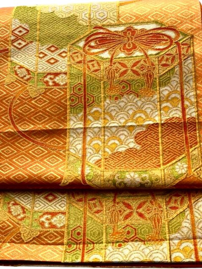Op de afbeelding: Detail van een gevouwen textiel met een ingewikkeld patroon. Het ontwerp bevat geometrische vormen, bloemmotieven en een grote strik in oranje, goud, groen en wit. De stof lijkt zijde of een vergelijkbaar luxueus materiaal te zijn.
