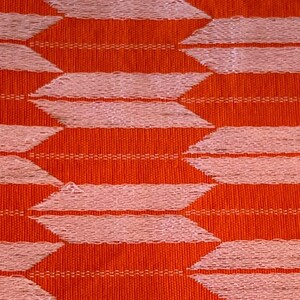May include: A woven fabric featuring a repeating geometric pattern of light pink, horizontal, arrow-like shapes arranged in staggered rows against a vibrant, solid orange background. The texture shows visible vertical and horizontal thread weaves.
