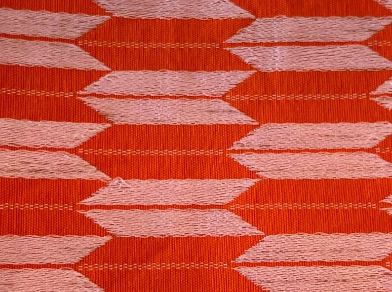 May include: A woven fabric featuring a repeating geometric pattern of light pink, horizontal, arrow-like shapes arranged in staggered rows against a vibrant, solid orange background. The texture shows visible vertical and horizontal thread weaves.