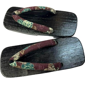 May include: A pair of traditional Japanese geta sandals. The wooden platforms are dark brown with a textured grain. The straps are burgundy with a floral pattern in shades of green and cream. These sandals are designed for outdoor wear.