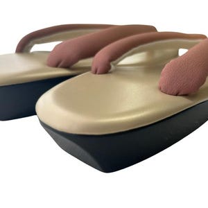 May include: A pair of traditional Japanese geta sandals. The sandals feature a beige platform with a black base and a pink thong strap. The design is simple and elegant, suitable for casual wear.
