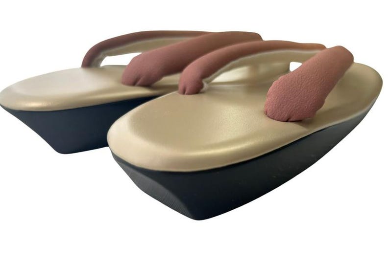 May include: A pair of traditional Japanese geta sandals. The sandals feature a beige platform with a black base and a pink thong strap. The design is simple and elegant, suitable for casual wear.