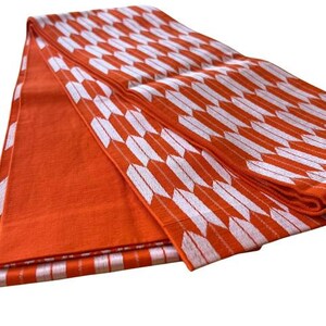 May include: A folded textile fabric featuring a vibrant orange base color with a repeating white arrow feather pattern. The material displays a textured weave, with one section showing a solid orange surface and another section showcasing the arrow motif.