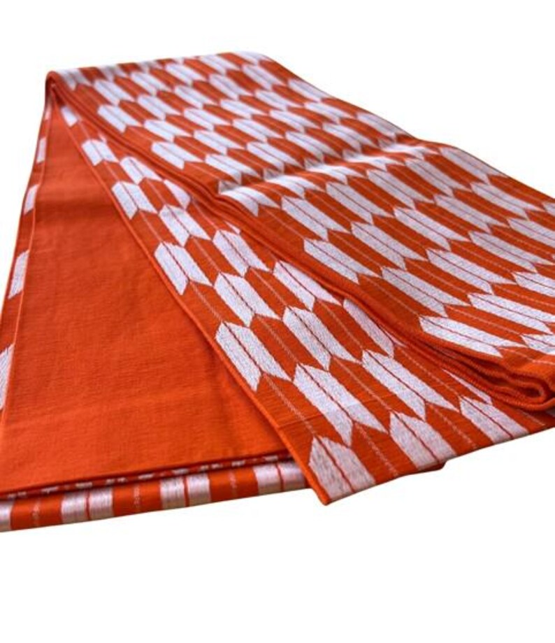 May include: A folded textile fabric featuring a vibrant orange base color with a repeating white arrow feather pattern. The material displays a textured weave, with one section showing a solid orange surface and another section showcasing the arrow motif.