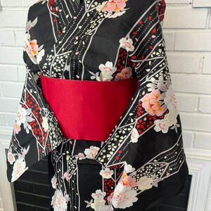 May include: A black kimono featuring a floral pattern with white and pink blossoms, accented by diagonal white and red stripes. The garment is cinched at the waist with a wide, solid red obi sash, creating a structured silhouette on a dress form.