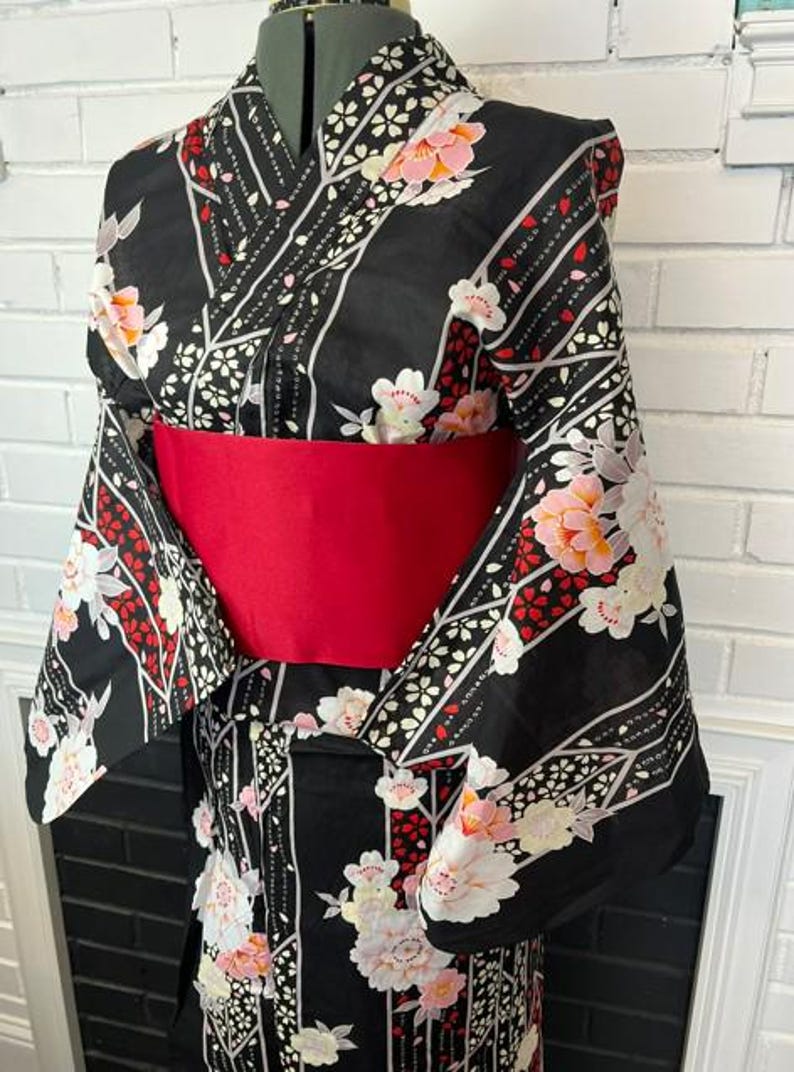 May include: A black kimono featuring a floral pattern with white and pink blossoms, accented by diagonal white and red stripes. The garment is cinched at the waist with a wide, solid red obi sash, creating a structured silhouette on a dress form.