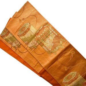 Op de afbeelding: Een oranje en gouden textiel met ingewikkelde geometrische en bloemmotieven. De stof heeft rechthoekige en ruitvormige vormen, met groene en bruine accenten. Het textiel is gevouwen en toont een effen oranje onderkant.