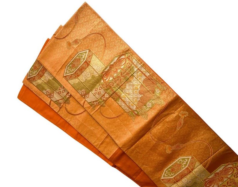 Op de afbeelding: Een oranje en gouden textiel met ingewikkelde geometrische en bloemmotieven. De stof heeft rechthoekige en ruitvormige vormen, met groene en bruine accenten. Het textiel is gevouwen en toont een effen oranje onderkant.