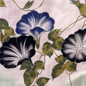 Op de afbeelding: Een stof met een botanische print van ochtendgloriebloemen in tinten diepblauw, paars en wit, vergezeld door olijfgroene bladeren en dunne, kronkelende wijnstokken op een bleke, lichtgekleurde achtergrond met een subtiel kleurverloop.