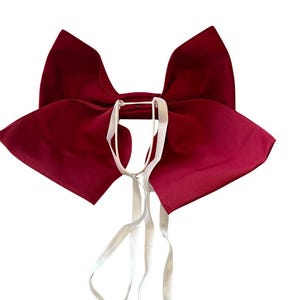 May include: A large, deep red fabric bow featuring two prominent, wide wings and a central knot. Attached to the center of the bow are two thin, white elastic straps that hang downward, designed for securing the accessory to a garment or item.