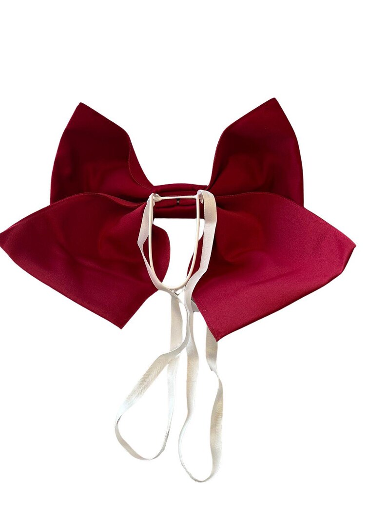 May include: A large, deep red fabric bow featuring two prominent, wide wings and a central knot. Attached to the center of the bow are two thin, white elastic straps that hang downward, designed for securing the accessory to a garment or item.
