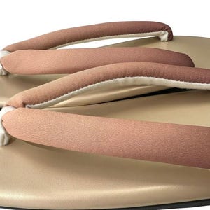 May include: Close-up of traditional Japanese geta sandals. The sandals feature a light tan base with a black sole and a soft, padded strap in shades of brown and white. The design is simple and elegant.