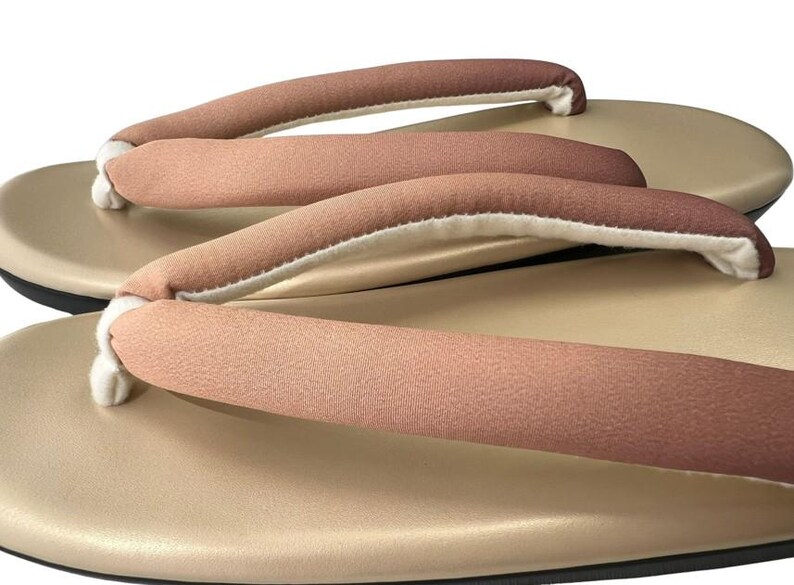 May include: Close-up of traditional Japanese geta sandals. The sandals feature a light tan base with a black sole and a soft, padded strap in shades of brown and white. The design is simple and elegant.