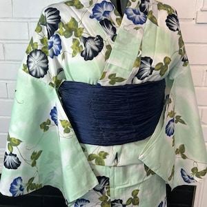 Op de afbeelding: Een licht muntgroene kimono met een herhalend patroon van blauwe en zwarte ochtendgloriebloemen met groene bladeren. Het kledingstuk is in de taille ingesnoerd met een brede, getextureerde, donker marineblauwe obi-sjerp.