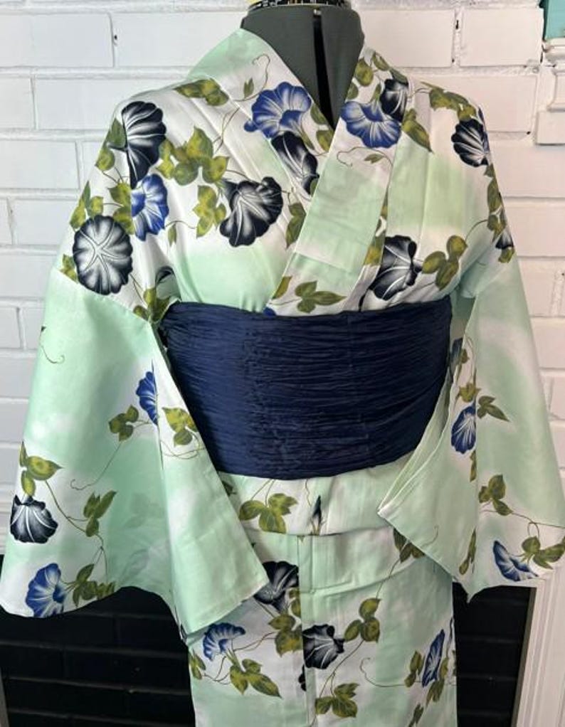 Op de afbeelding: Een licht muntgroene kimono met een herhalend patroon van blauwe en zwarte ochtendgloriebloemen met groene bladeren. Het kledingstuk is in de taille ingesnoerd met een brede, getextureerde, donker marineblauwe obi-sjerp.