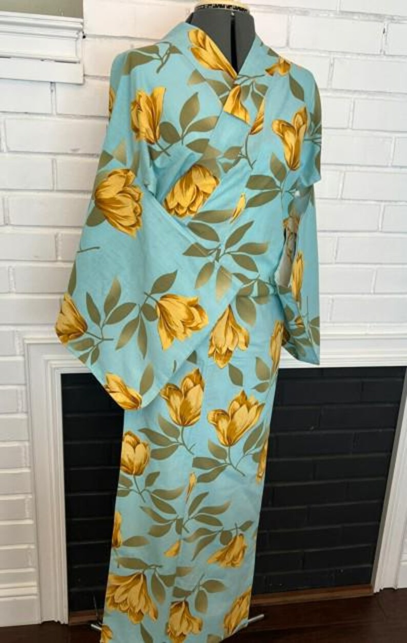 May include: A light blue kimono featuring a repeating pattern of golden yellow tulip-like flowers and muted green leaves. The garment has wide sleeves, a wrap-front closure, and a structured collar, displayed on a mannequin torso.