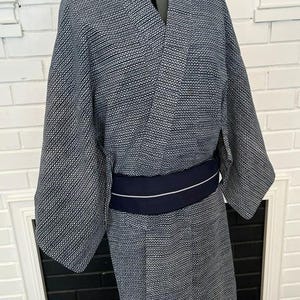 Może przedstawiać: Granatowo-białe kimono we wzory o drobnej geometrycznej fakturze na całej powierzchni tkaniny. Ubi&oacute;r posiada szerokie rękawy i kopertowy prz&oacute;d, przewiązany w talii jednolitym, ciemnogranatowym pasem obi z cienkim białym paskiem.