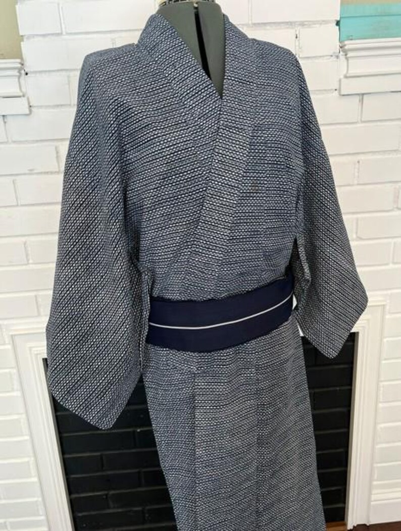 Może przedstawiać: Granatowo-białe kimono we wzory o drobnej geometrycznej fakturze na całej powierzchni tkaniny. Ubi&oacute;r posiada szerokie rękawy i kopertowy prz&oacute;d, przewiązany w talii jednolitym, ciemnogranatowym pasem obi z cienkim białym paskiem.