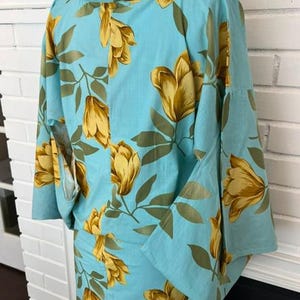 May include: A light blue kimono featuring a repeating pattern of large golden-yellow flowers and green leaves. The garment has a high collar, wide sleeves, and a straight-cut silhouette, displayed on a mannequin to show the floral print design.