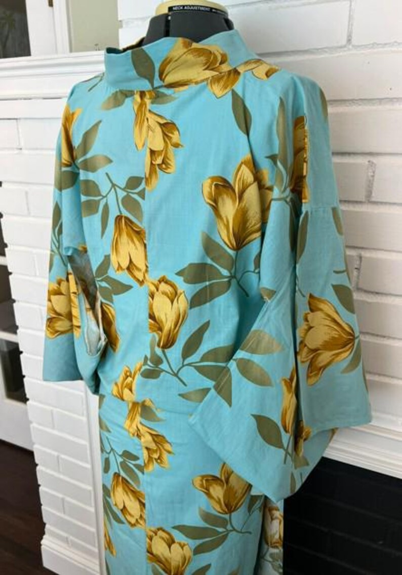 May include: A light blue kimono featuring a repeating pattern of large golden-yellow flowers and green leaves. The garment has a high collar, wide sleeves, and a straight-cut silhouette, displayed on a mannequin to show the floral print design.