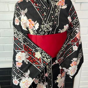 May include: A black kimono featuring a floral pattern with white, pink, and red blossoms, accented by geometric lines and small petal motifs. The garment is cinched at the waist with a wide, solid red obi sash, creating a structured silhouette.