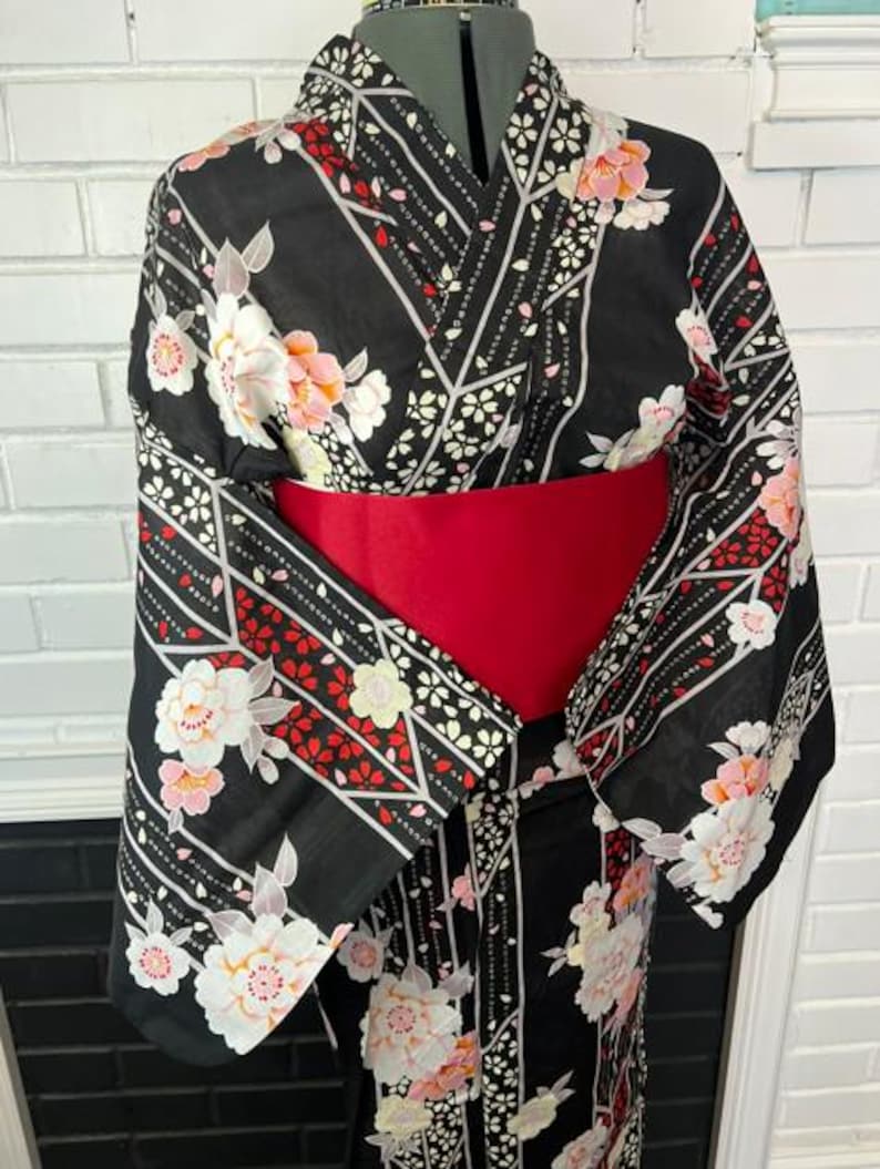 May include: A black kimono featuring a floral pattern with white, pink, and red blossoms, accented by geometric lines and small petal motifs. The garment is cinched at the waist with a wide, solid red obi sash, creating a structured silhouette.