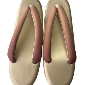May include: A pair of beige Japanese geta sandals. The sandals have a rounded shape with a light brown strap that is a darker shade at the bottom. The sandals are made of a smooth material.