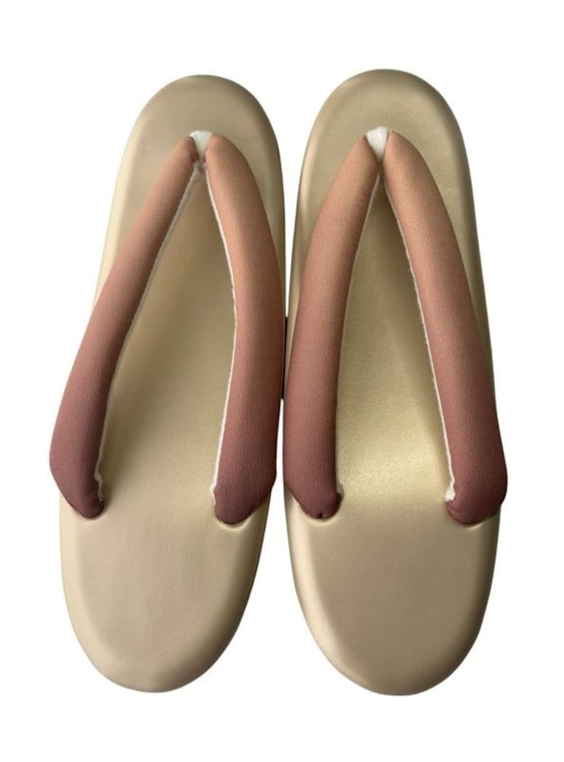 May include: A pair of beige Japanese geta sandals. The sandals have a rounded shape with a light brown strap that is a darker shade at the bottom. The sandals are made of a smooth material.